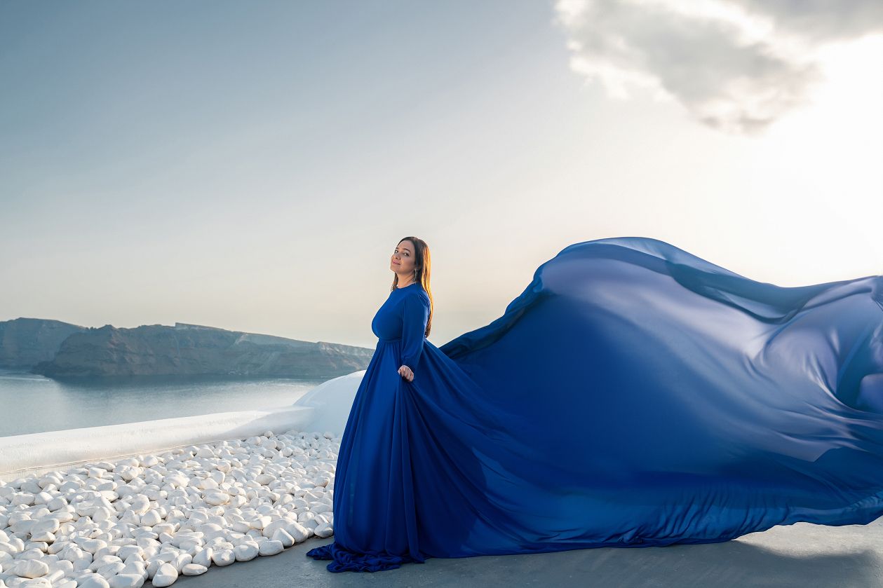 A royal blue long sleeve flying dress billows magnificently over Santorini's white pebble terrace as the caldera cliffs fade into a dreamy golden horizon.